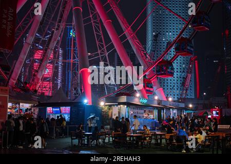 Hong Kong, Cina - 2 aprile 2023: Folla di persone sotto la ruota panoramica nel fronte del porto con luci notturne urbane Foto Stock