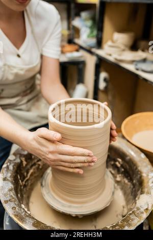 Vista a ritaglio di una donna di vasaio che scompone l'argilla sulla ruota della ceramica in un laboratorio di ceramica sfocato Foto Stock