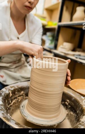 Vista ritagliata dell'artigiana femminile in grembiule che forma l'argilla su ruota in ceramica in officina sfocata Foto Stock