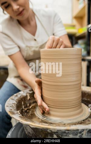 Vista ritagliata di una donna sfocata che crea un vaso di argilla su una ruota in ceramica in officina sullo sfondo Foto Stock