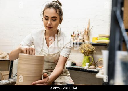 Bruna artigiana in grembiule che produce vaso in argilla e lavora su una ruota in ceramica in un laboratorio di ceramica Foto Stock