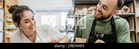 Ceramisti gioiosi in grembiuli che muoiono in una ciotola di argilla insieme mentre lavoravano in uno studio di ceramica sullo sfondo Foto Stock