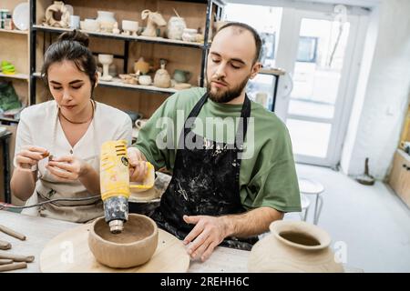 Un paio di artigiani in grembiuli che lavorano insieme con pistola termica e argilla in uno studio di ceramica sullo sfondo Foto Stock