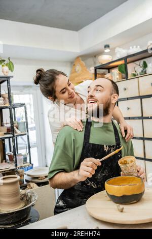 Artigiana positiva in grembiule che abbraccia e guarda la ciotola di argilla colorata fidanzata nello studio di ceramica Foto Stock