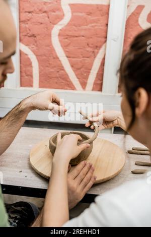 Coppia di artigiani sfocati che modellano la ciotola di argilla su tavola di legno nello studio di ceramica sullo sfondo Foto Stock