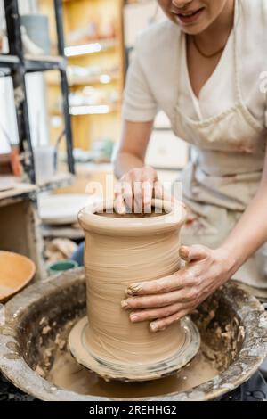 Vista parziale della donna in vaso di argilla sfocata che forma il grembiule sulla ruota in ceramica nello studio di ceramica Foto Stock