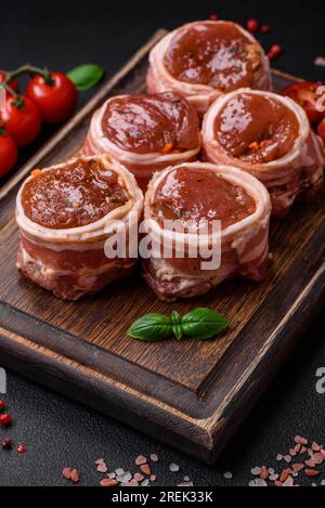 Deliziosi involtini di carne di maiale o di pollo crudi avvolti in pancetta, con sale, spezie ed erbe su fondo di cemento scuro Foto Stock