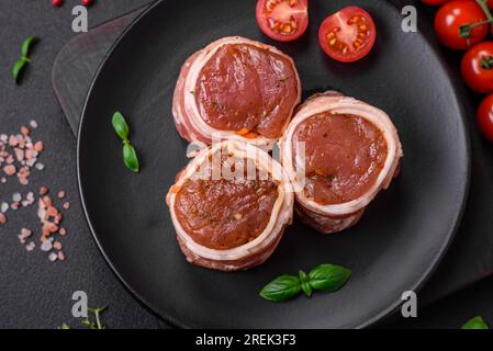 Deliziosi involtini di carne di maiale o di pollo crudi avvolti in pancetta, con sale, spezie ed erbe su fondo di cemento scuro Foto Stock