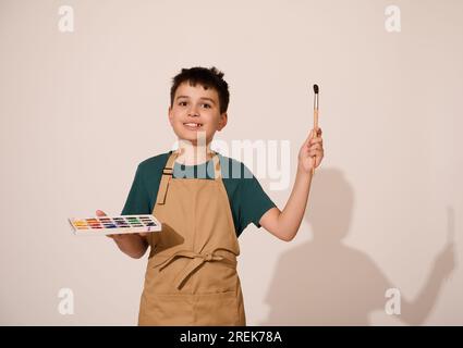 Adorabile bambino che si diverte a lezione d'arte, puntando un pennello su uno spazio copia, tenendo in mano una tavolozza di colorate e vivaci pitture ad acquerello, isolate sopra W. Foto Stock