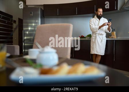 Il maschio barbuto si trova nella zona cucina con il telefono cellulare Foto Stock