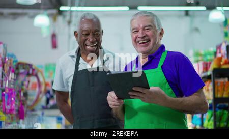 Gioioso e diversificato personale brasiliano della catena di supermercati sorridente alla macchina fotografica con tavolo e uniformi. Afroamericano anziano impiegato e una CA Foto Stock