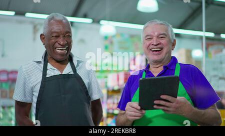 Gioioso e diversificato personale brasiliano della catena di supermercati sorridente alla macchina fotografica con tavolo e uniformi. Afroamericano anziano impiegato e una CA Foto Stock