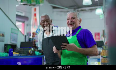 Gioioso e diversificato personale brasiliano della catena di supermercati sorridente alla macchina fotografica con tavolo e uniformi. Afroamericano anziano impiegato e una CA Foto Stock