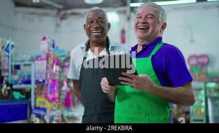 Gioioso e diversificato personale brasiliano della catena di supermercati sorridente alla macchina fotografica con tavolo e uniformi. Afroamericano anziano impiegato e una CA Foto Stock