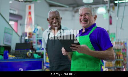 Gioioso e diversificato personale brasiliano della catena di supermercati sorridente alla macchina fotografica con tavolo e uniformi. Afroamericano anziano impiegato e una CA Foto Stock