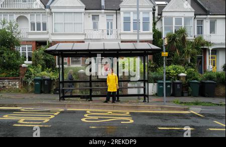 Una donna con un cappotto giallo brillante in attesa di un autobus presso una fermata dell'autobus nell'area di Preston Park a Brighton. 27 luglio 2023 Foto Stock