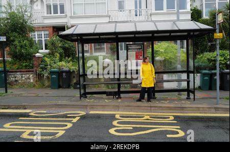 Una donna con un cappotto giallo brillante in attesa di un autobus presso una fermata dell'autobus nell'area di Preston Park a Brighton. 27 luglio 2023 Foto Stock