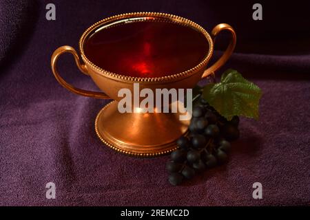 Calice di colore dorato con liquido rosso e uva accanto su un panno viola Foto Stock