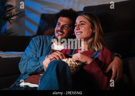 Giovane coppia caucasica sorridente e divertita a guardare programmi televisivi o film a tarda notte seduti sul divano a casa a mangiare popcorn. Due amici che si divertono con il servizio di streaming su uno smart tv. Foto di alta qualità Foto Stock
