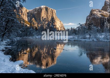 La superficie rocciosa delle montagne della Sierra Nevada, che riflette la luce del sole del mattino presto, si riflette nel fiume Merced, in un paesaggio innevato di inizio inverno nel parco nazionale di Yosemite. Foto Stock