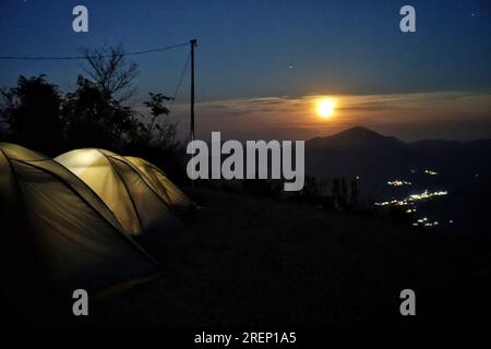 Campi illuminati su un campeggio con una luna piena che si alza su una collina sullo sfondo. Trekking e campeggio nell'Himalaya. Uttarakhand India. Foto Stock