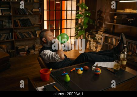 Uomo d'affari anziano e rilassato che fa saltare in aria la mongolfiera mentre è seduto in ufficio Foto Stock