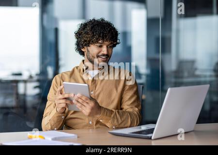Giovane ispanico di successo con un tablet, soddisfatto dei risultati ottenuti sorridendo guardando lo schermo, uomo che usa un'applicazione, seduto sul posto di lavoro con un notebook all'interno dell'ufficio. Foto Stock