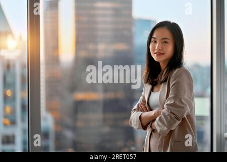 Giovane donna d'affari asiatica sicura di sé, leader in piedi, ha attraversato le braccia alla finestra. Foto Stock