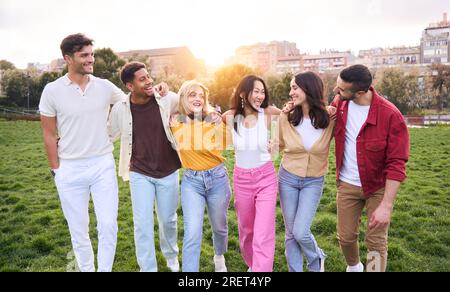 Giovane attraente gruppo multirazziale che cammina verso la fotocamera con il tramonto dietro di loro che calpestano l'erba Foto Stock
