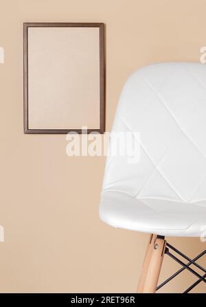 sedia bianca trapuntata vicino alla parete beige, cornice marrone con carta kraft appesa alla parete - spazio per il testo, vista frontale, spazio per le copie, mockup Foto Stock