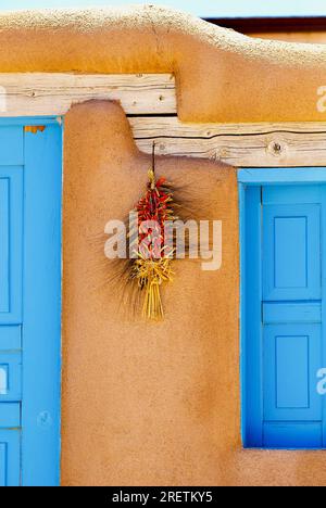 Ranchos de Taos, New Mexico, USA - 23 luglio 2023: Una ristra è appesa accanto a una serranda di legno dipinta di blu sulla parete esterna di una tradizionale casa di adobe. Foto Stock