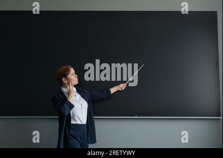Donna caucasica dai capelli rossi con un vestito da pantaloni. Insegnante premurosa con un puntatore alla lavagna. Foto Stock