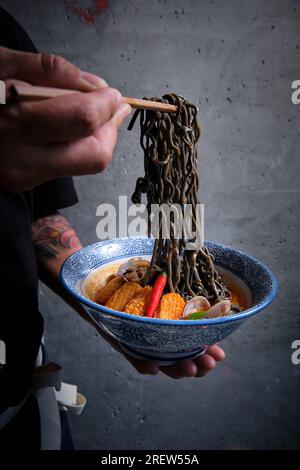 Mano maschile raccogliendo tagliatelle nere in una ciotola di ramen caldo con brodo di pesce, calamari, peperoncini, vongole. Foto Stock