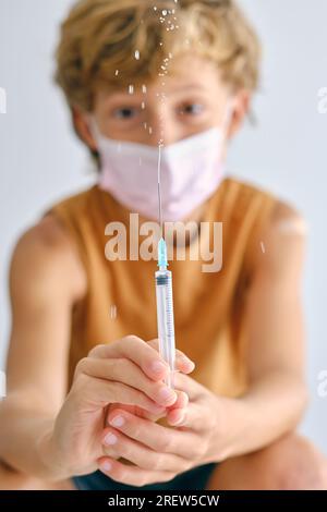 Bambino in maschera che guarda la macchina fotografica mentre spruzza il vaccino dall'iniettore durante la pandemia di COVID 19 su sfondo chiaro Foto Stock