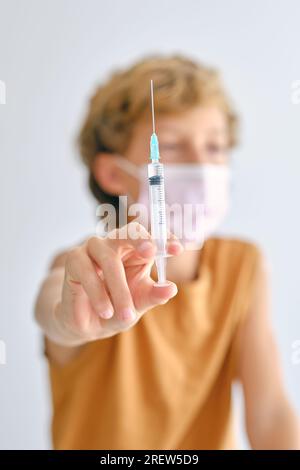 Bambino in maschera sterile che mostra iniettore con medicina liquida mentre guarda lontano durante la pandemia di COVID 19 su sfondo chiaro Foto Stock