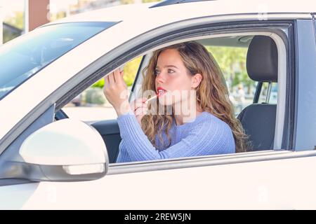 Bella giovane driver femminile in pullover blu che applica rossetto rosso brillante e guardando in visiera parasole mentre si siede in auto bianca in giornata di sole Foto Stock