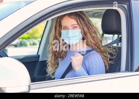 Giovane e attraente conducente donna che indossa maschera facciale e pullover blu seduto sul sedile del conducente dell'auto con cintura allacciata e guardando la telecamera Foto Stock