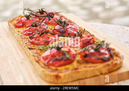 Pane piatto di forma rettangolare con fette di pomodoro e ciuffi di timo su tagliere di legno su sfondo sfocato Foto Stock