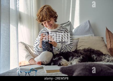 Allegro scolaro con fotocamera vintage seduto con gambe incrociate vicino Miniature Schnauzer sul letto in casa Foto Stock