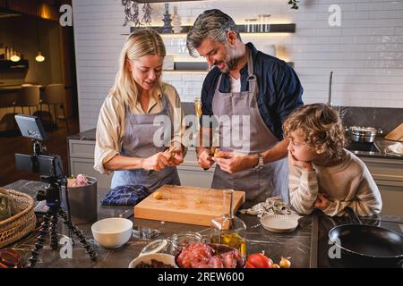 Simpatico ragazzo che guarda i genitori che sbucciano le patate crude al tavolo e registrano il vlog mentre cucinano insieme in una moderna cucina leggera Foto Stock