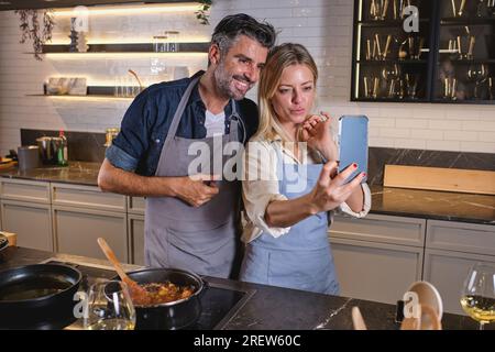 Coppia multirazziale positiva in grembiuli in piedi vicino alla stufa elettrica e scattando un autoritratto sul telefono cellulare mentre cucinano il cibo insieme a casa Foto Stock