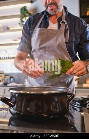 Chef maschio professionista in piedi al piano cottura e tenendo un mazzo di foglie di alloro mentre prepari una deliziosa zuppa nella cucina moderna a casa Foto Stock