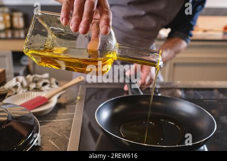 Tagliare il cuoco maschile anonimo versando l'olio dalla bottiglia nella padella sul piano cottura mentre si cucina al bancone in una cucina leggera Foto Stock