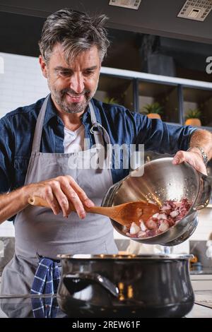 Uno chef maschio positivo in grembiule in piedi al piano cottura e versare il polpo nella pentola mentre si cucina in cucina a casa Foto Stock