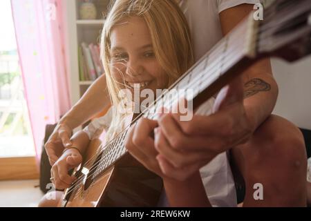 Ragazza bionda che suona la chitarra mentre sorride con una donna seduta con lei su un divano e insegnandola in un casa Foto Stock