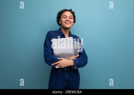 una giovane e snella bruna ispanica con morbidi capelli ricci in un tuta di denim blu può contenere documenti e un computer portatile per il lavoro Foto Stock