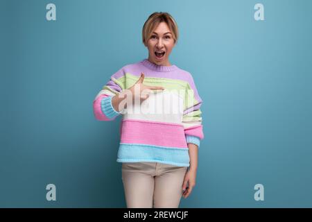 una giovane donna bionda sorpresa con un abito casual che punta al muro con un dito su sfondo blu e uno spazio per le copie Foto Stock