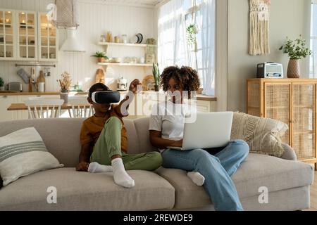 Moderna famiglia afro-americana, mamma e figlio si siedono sul divano con dispositivi elettronici. Tecnologia e bambini Foto Stock