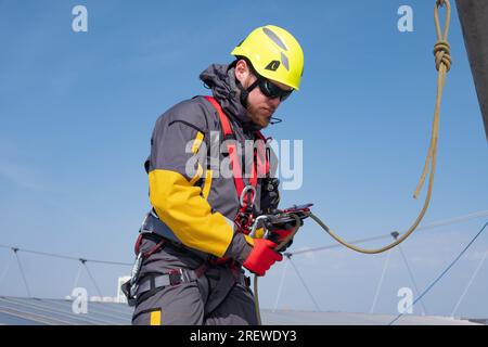 Alpinista industriale maschio che fissa una fune a un dispositivo di chiusura sul tetto di un cantiere edile, preparandosi al lavoro. Un lavoratore che indossa una cintura di sicurezza e un casco lavora in altezza osservando le precauzioni di sicurezza Foto Stock