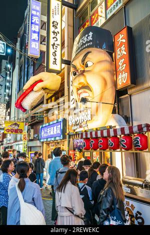 La gente fa la coda di notte fuori dall'entrata del popolare ristorante Kushikatsu daruma con la scultura arrabbiata dello chef sopra la testa a Dotonbori, Osaka. Foto Stock